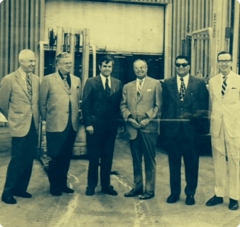 Photograph of six men in suits standing in front of a warehouse.